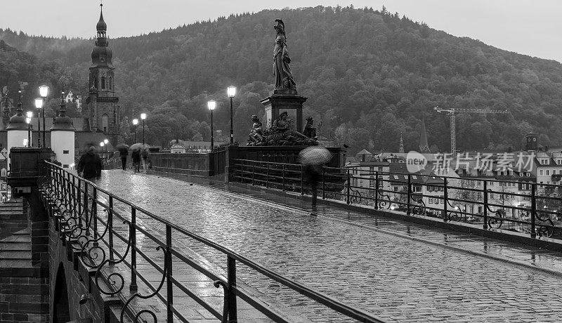在一个下雨天，海德堡的老桥(karl - theodor - brcke)的黑白照片。一些行人打着伞走着。模糊了桥上人群的移动效果。